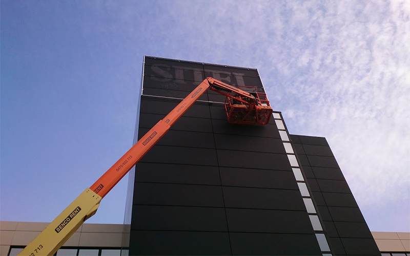 Een medewerker van All Signs staat op een kraan om een spandoekframe aan een toren te hangen.